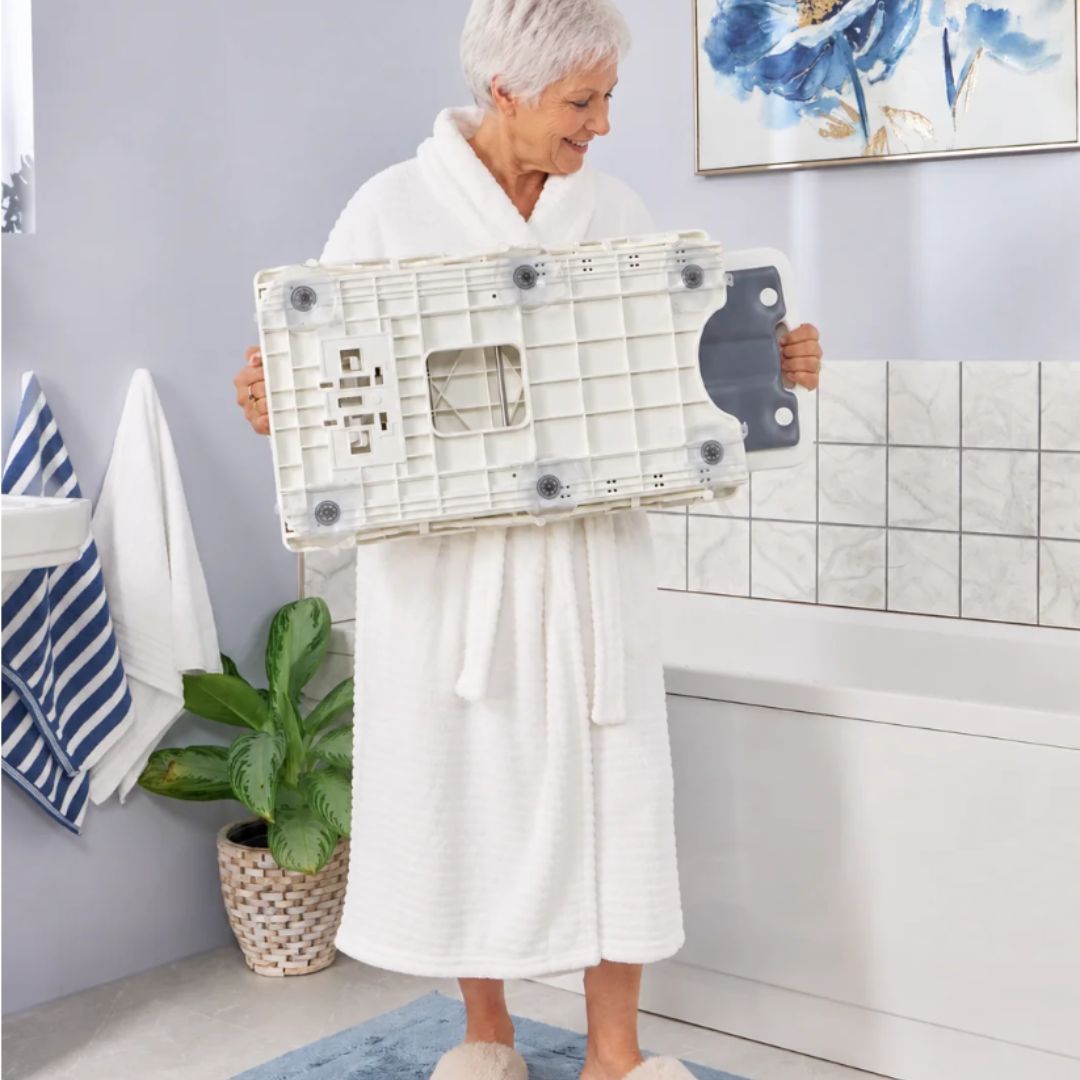 An older woman in a white bathrobe stands in a bathroom holding a white NRS riverdale folded bath lift with gray non-slip pads and a dark gray handle.