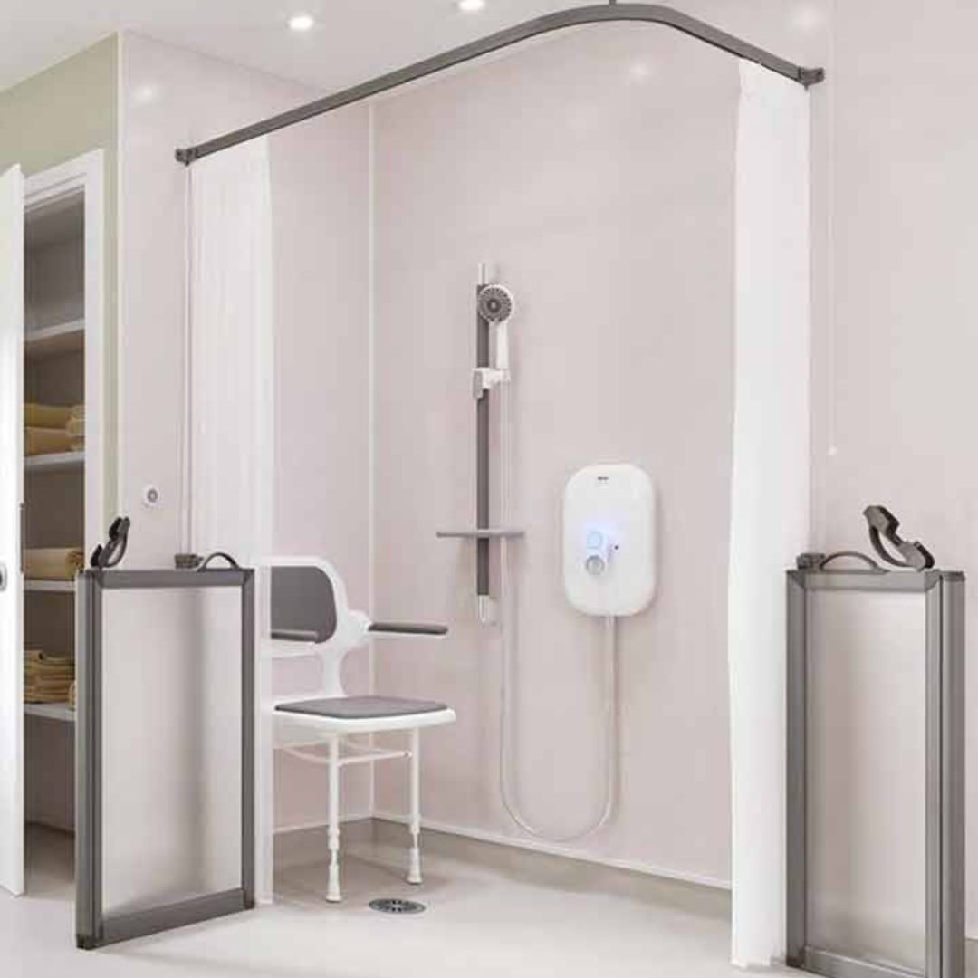 A modern shower bench setup in a bathroom, featuring a white shower chair with armrests, a wall-mounted shower head and hose, a white electronic water heater, white curtains, and gray-framed shower doors and handrails.