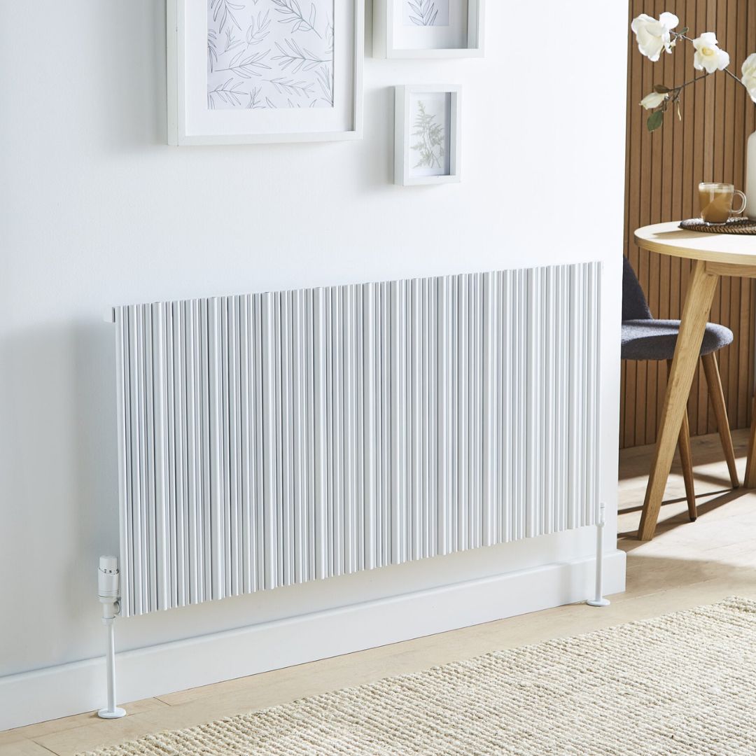 A modern white pleated radiator mounted on a white wall in a bright room, with framed botanical prints above and a wooden dining table and chair partially visible on the right.