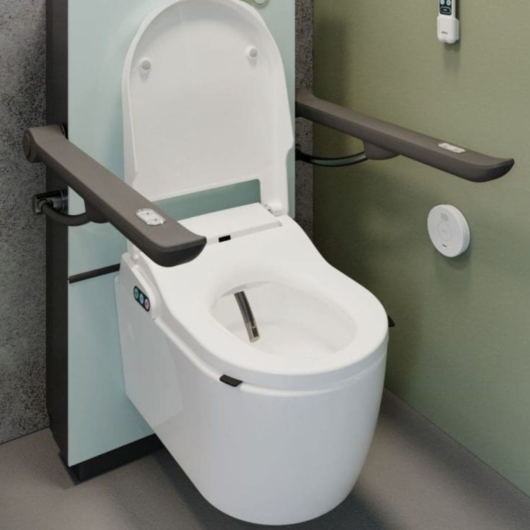 A white bidet toilet with an open seat and raised lid, fitted with dark gray grab handles mounted on both sides, shown in a bathroom setting with a pale green wall and gray floor.