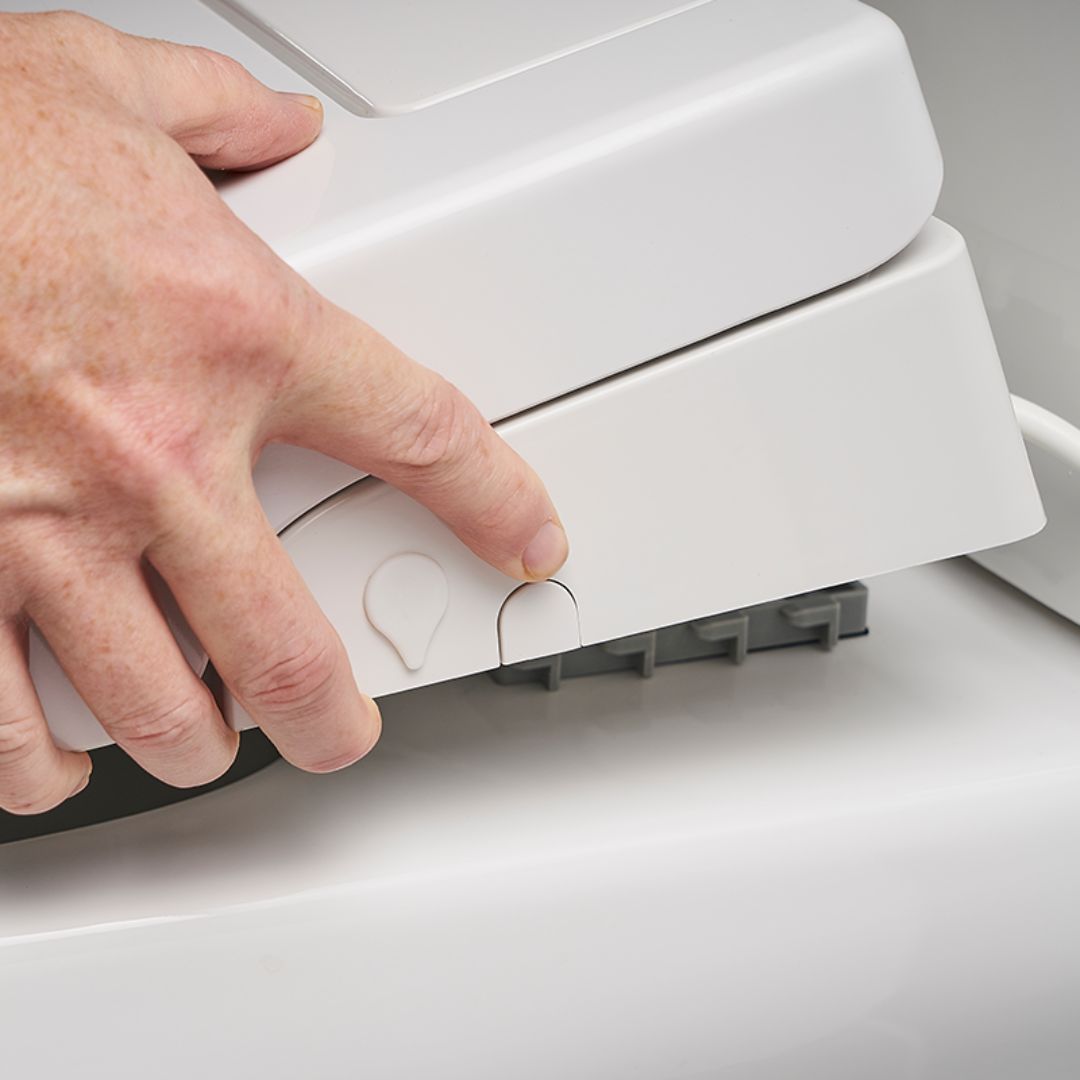A close-up of a person's hand pressing a small button on a white bidet toilet seat to remove