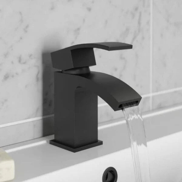 Black faucet on a bathroom sink with water flowing, against a light gray tiled wall.