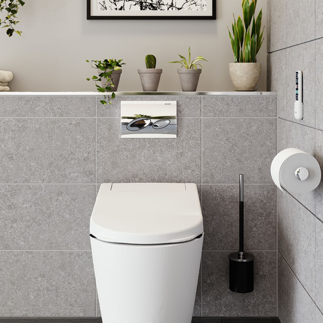 A modern white toilet with a flush button on the tiled wall, shown in a clean, contemporary bathroom setting with gray wall tiles, small potted plants, and a toilet brush holder.