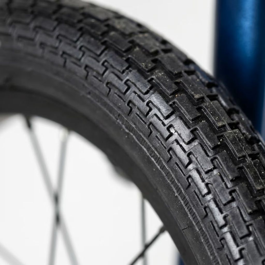 Close-up of a black wheelchair tyre with a zigzag tread pattern, mounted on a wheel with thin silver spokes, against a white background with a blue frame element visible on the right.