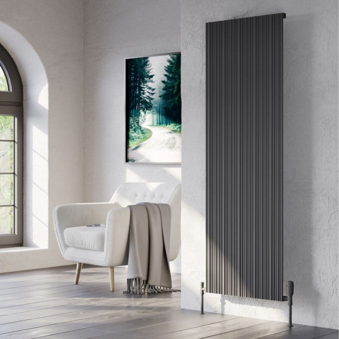 A tall black radiator with vertical fluted panels, mounted on a textured white wall in a modern living room setting next to a white armchair and a framed forest print.