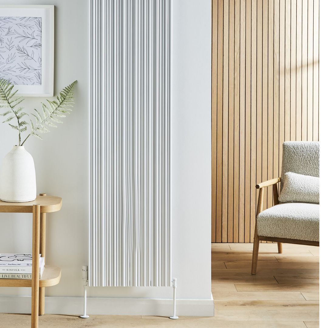 A tall, modern white radiator with vertical fluted slats, shown in a styled room setting with a light wood-paneled wall, a wooden side table with a white vase, and a textured armchair.