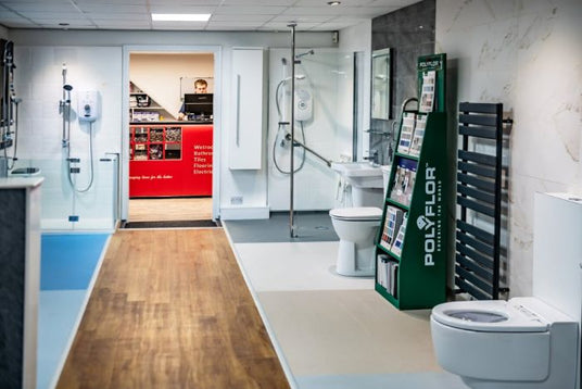 A showroom interior with multiple bathroom fixtures, including a white toilet, a freestanding bathtub, a shower area, and a green POLOFLOR wood floor display board in the foreground.