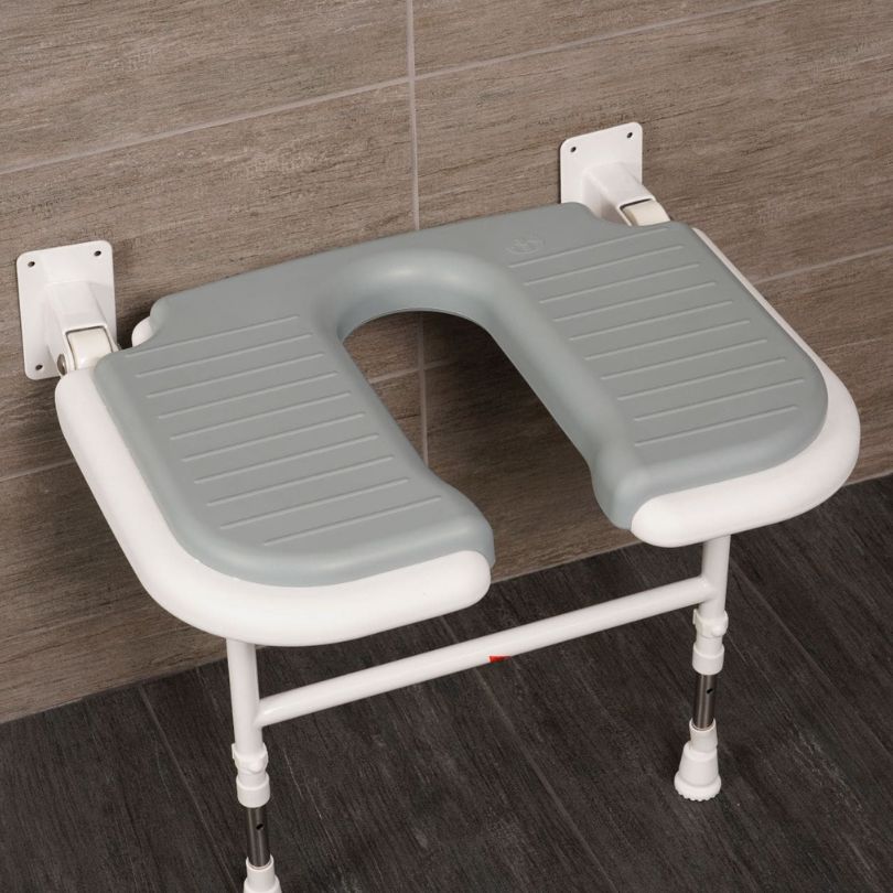 A wall-mounted bathroom horseshoe safety seat with a grey molded seat cushion and white frame, shown attached to a tiled wall in a bathroom setting.