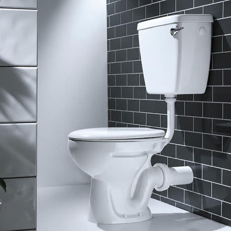 A white ceramic toilet with a wall-mounted cistern and white drainage pipe, set against a black subway tile wall and a white wall.