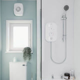 A modern bathroom scene showing a white wall-mounted shower valve and flexible shower hose on a white tiled wall, alongside a window, toilet, and small plant.