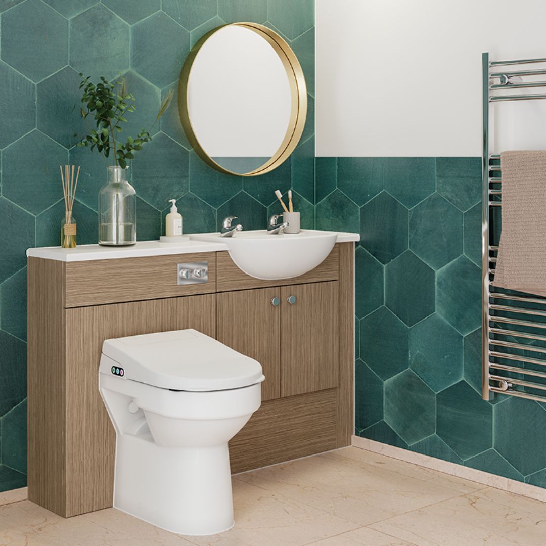 A modern bathroom vanity setup with a white sink and toilet, wood-look cabinet doors, a round gold-framed mirror, and teal hexagonal wall tiles. A towel rail with a beige towel is visible on the right.