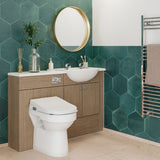 A modern bathroom vanity setup with a white sink and toilet, wood-look cabinet doors, a round gold-framed mirror, and teal hexagonal wall tiles. A towel rail with a beige towel is visible on the right.