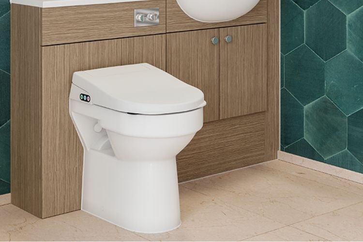 A white wall-mounted bidet toilet with a closed lid and a small control panel on the left side, installed in front of a wood-look vanity cabinet with a sink partially visible above it. The bathroom has green hexagonal wall tiles and light beige floor tiles.