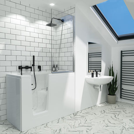 A modern white bathtub with a black shower fixture in a bright white-tiled bathroom. The room features a black-framed skylight, a white wall-mounted sink, a potted plant, and a patterned floor.