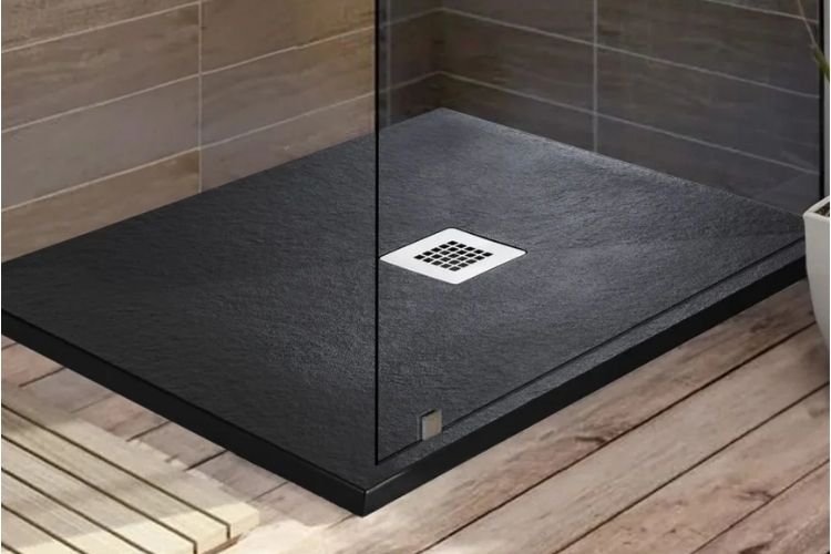 A modern minimalist shower floor in dark charcoal gray with a square white drain, shown in a bathroom setting with light wood flooring and beige wall tiles.