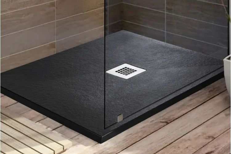 A modern square shower tray with a dark textured finish and a central square drain, shown with a clear glass door in a bathroom setting with wood-look flooring and tiled walls.