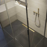 A modern walk-in shower enclosure with a clear glass door, black frame, and brass shower fixture on a light beige tiled wall and dark charcoal floor.