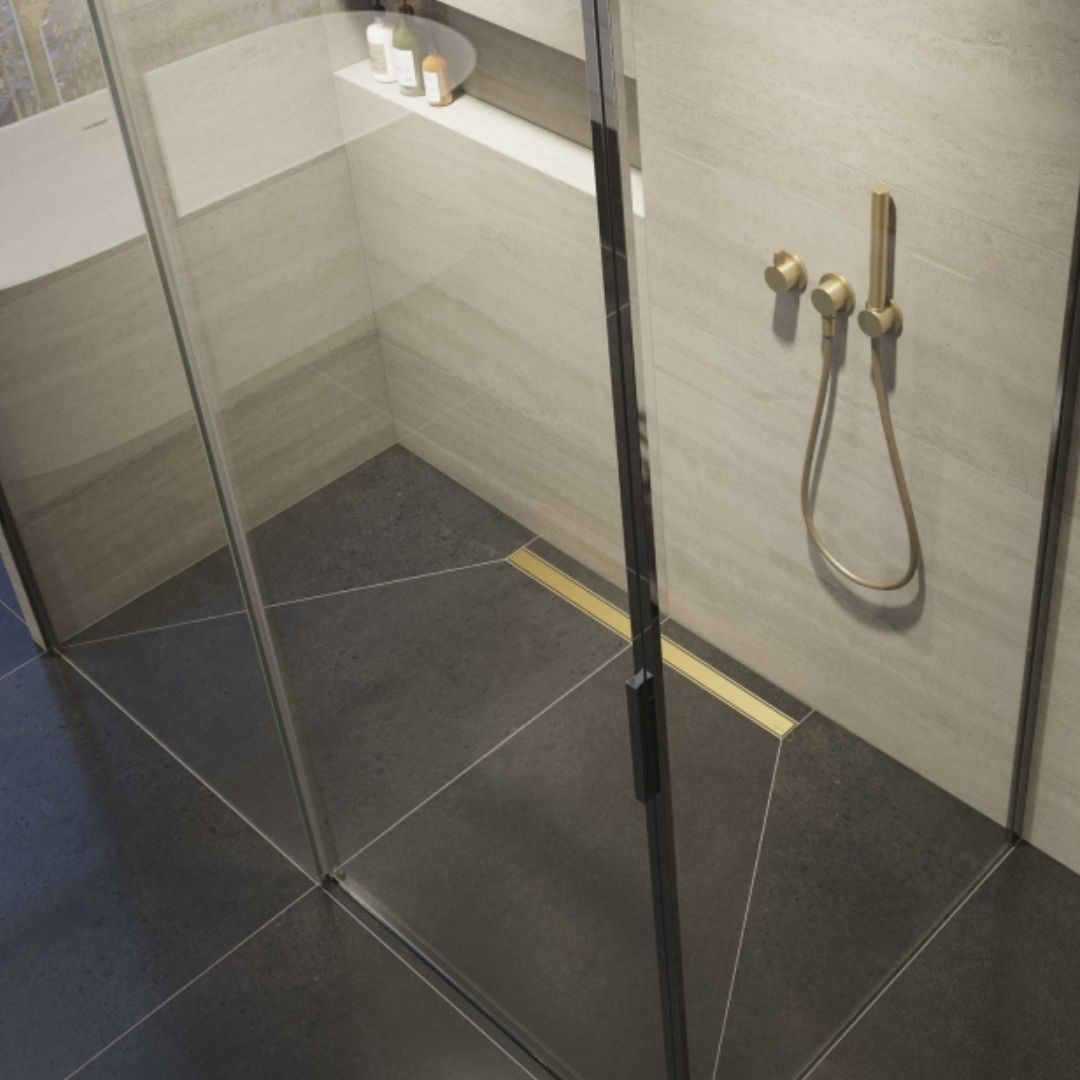 A modern walk-in shower enclosure with a clear glass door, black frame, and brass shower fixture on a light beige tiled wall and dark charcoal floor.