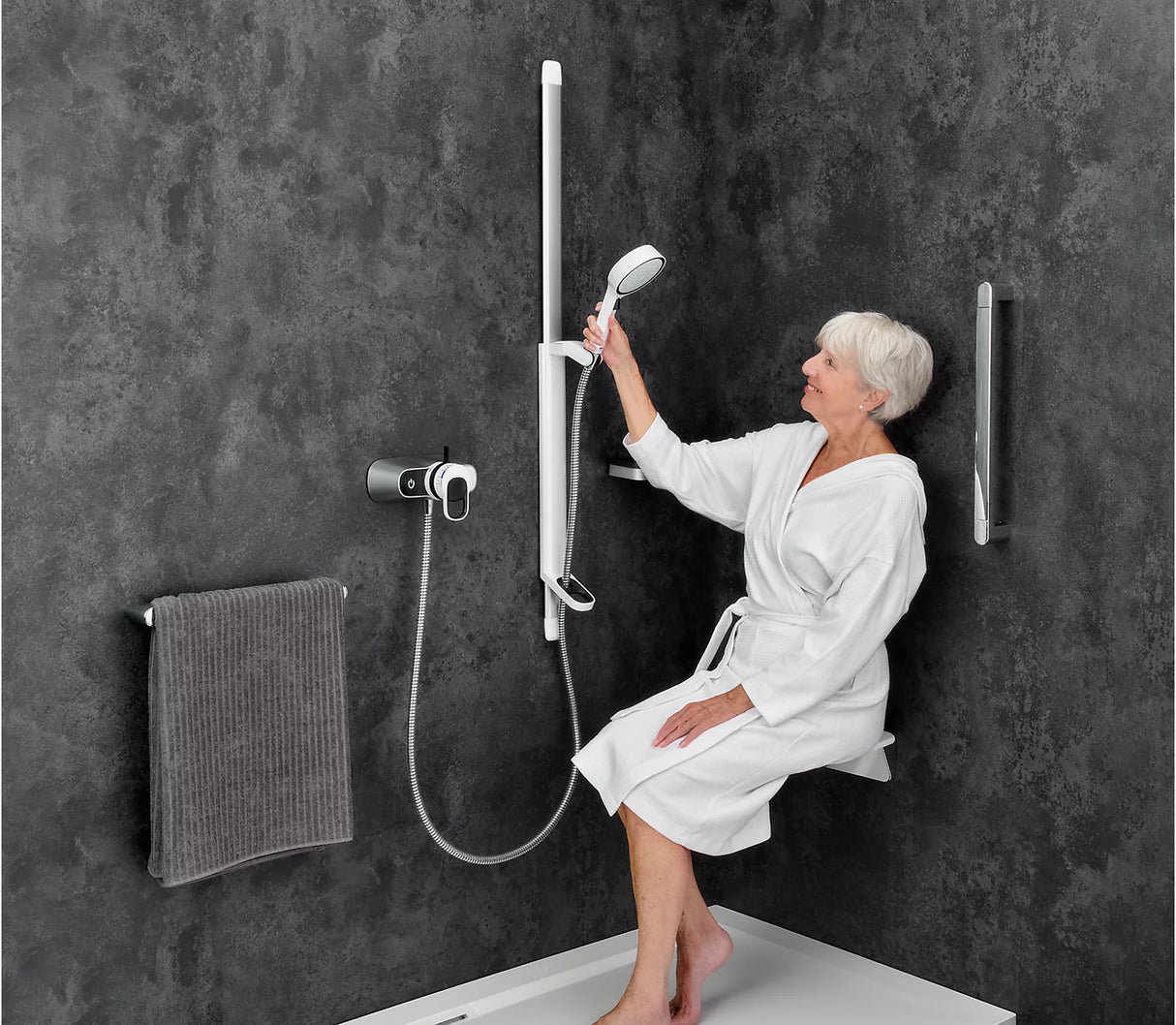Woman in a white robe using a shower head in a modern bathroom with dark walls.