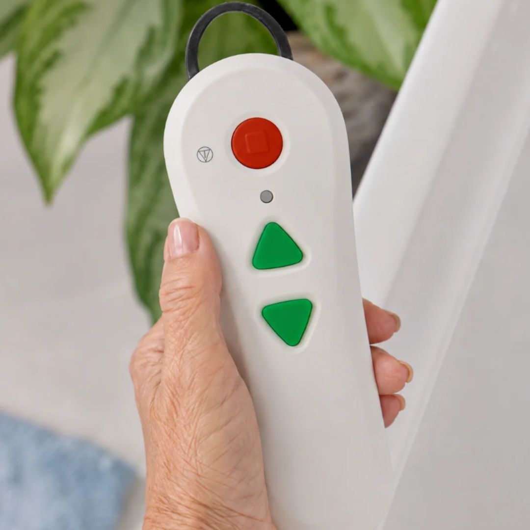 A hand holding a white handheld remote control with green arrow buttons, a red round button, and a black hanging loop, with a potted plant and white wall in the background.