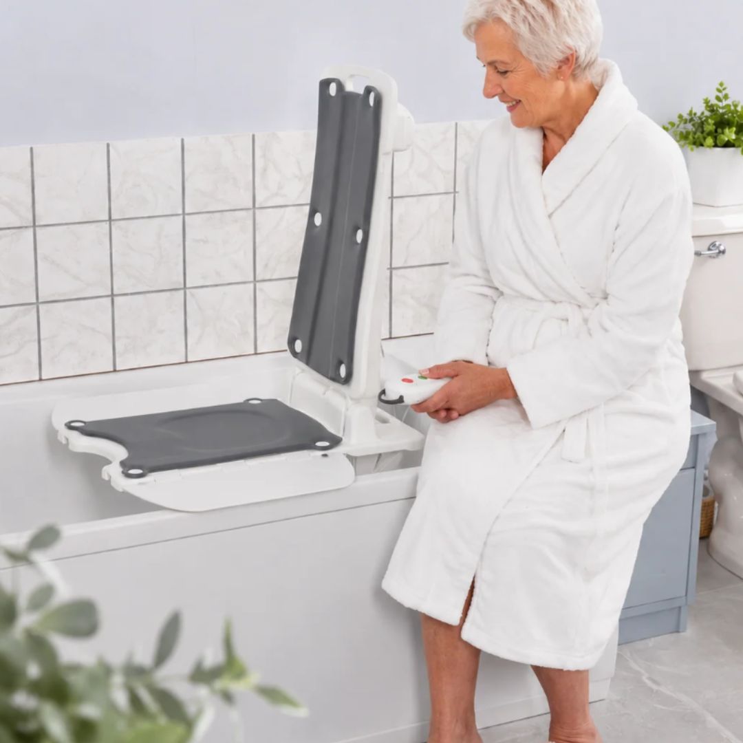 An older woman in a white bathrobe sits beside a bathtub in a tiled bathroom while using a white and grey raised bath lift.