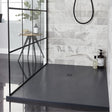 A modern shower area with a dark charcoal gray floor and matching dark countertop, set against a light gray marble-look tiled wall. A black-framed glass partition is visible on the left, and a small wooden side table with a bottle and plant sits inside the shower.