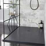 A modern shower area with a dark charcoal gray floor and matching dark countertop, set against a light gray marble-look tiled wall. A black-framed glass partition is visible on the left, and a small wooden side table with a bottle and plant sits inside the shower.