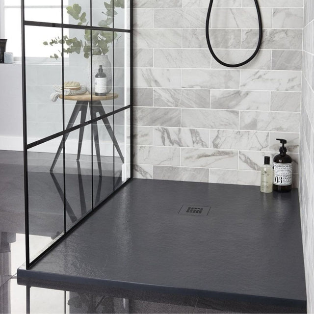 A modern shower area with a dark charcoal gray floor and matching dark countertop, set against a light gray marble-look tiled wall. A black-framed glass partition is visible on the left, and a small wooden side table with a bottle and plant sits inside the shower.