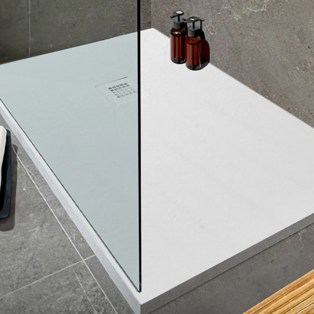 A modern white rectangular shower base with a clear glass panel and gray tiled bathroom walls and floor, with two dark amber pump bottles placed on the glass.