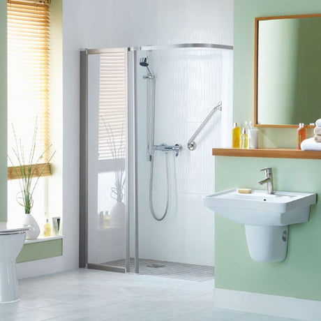 A modern corner shower enclosure with a glass door and silver frame, installed in a bright bathroom with a white sink, pale green walls, and a window with blinds.