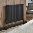 A black ribbed panel radiator with support feet, mounted on a wood-paneled wall in a modern living room setting.
