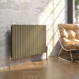 A modern horizontal panel radiator in a warm metallic brass finish, styled in a room with a distressed plaster wall, light wood flooring, and a yellow leather armchair.