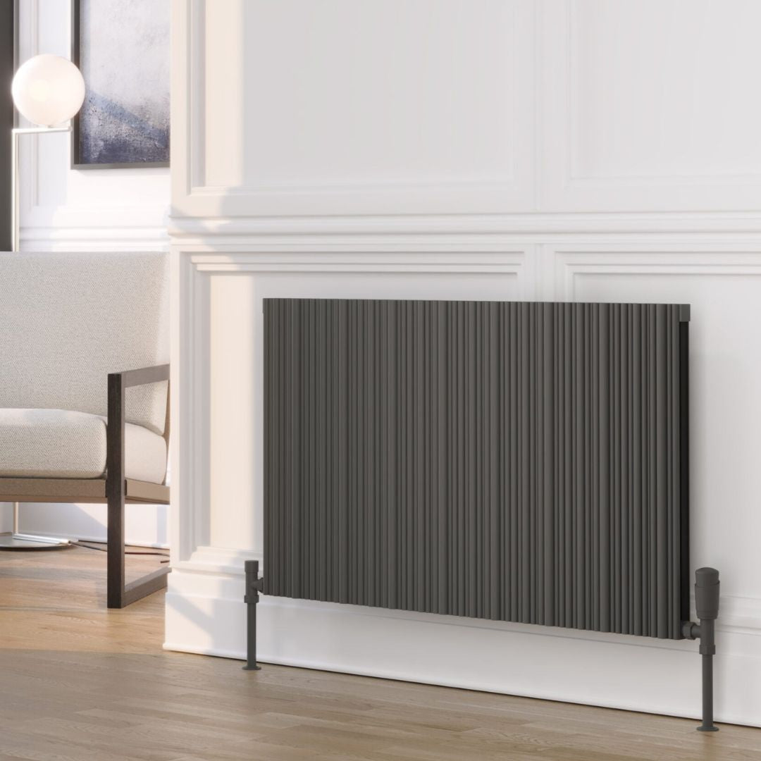 A dark gray pleated panel radiator shown in a modern living room setting with a white paneled wall, light wood floor, and a partial view of a white armchair.