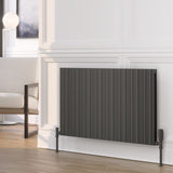 A dark gray pleated panel radiator shown in a modern living room setting with a white paneled wall, light wood floor, and a partial view of a white armchair.