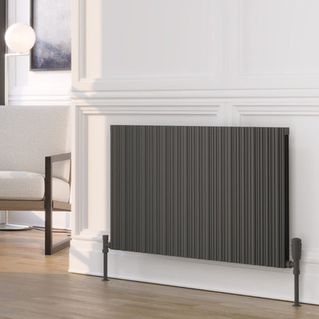 A dark gray pleated panel radiator shown in a modern living room setting with a white paneled wall, light wood floor, and a partial view of a white armchair.