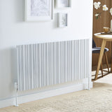 A modern white pleated radiator mounted on a white wall in a bright room, with framed botanical prints above and a wooden dining table and chair partially visible on the right.