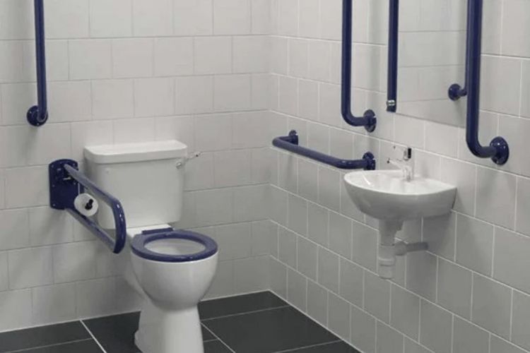 Accessible bathroom with white tiled walls, a white toilet with blue grab handles, a white wall-mounted sink with blue grab handles, and a dark gray floor.