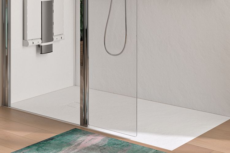 A modern frameless glass shower door installed in a minimalist white walk-in shower with a white shower tray, chrome hand shower, and light wood flooring. A green and pink abstract patterned bath mat is visible in the foreground.