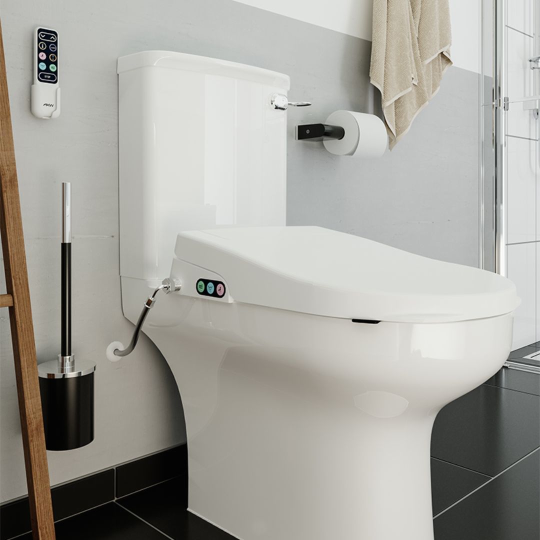 A white bidet toilet with a closed lid and seat, shown installed in a modern bathroom. The toilet has small colored indicator lights on the front, a connected bidet sprayer hose, a wall-mounted toilet paper holder, and a remote control mounted on the wall.