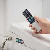 A hand holding a white remote control with a black touch panel, demonstrating its use on a white bidet toilet seat with additional control buttons visible on the side.