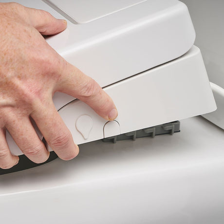 A close-up of a person's hand pressing a small button on a white bidet toilet seat to remove