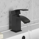 Black faucet on a bathroom sink with water flowing, against a light gray tiled wall.