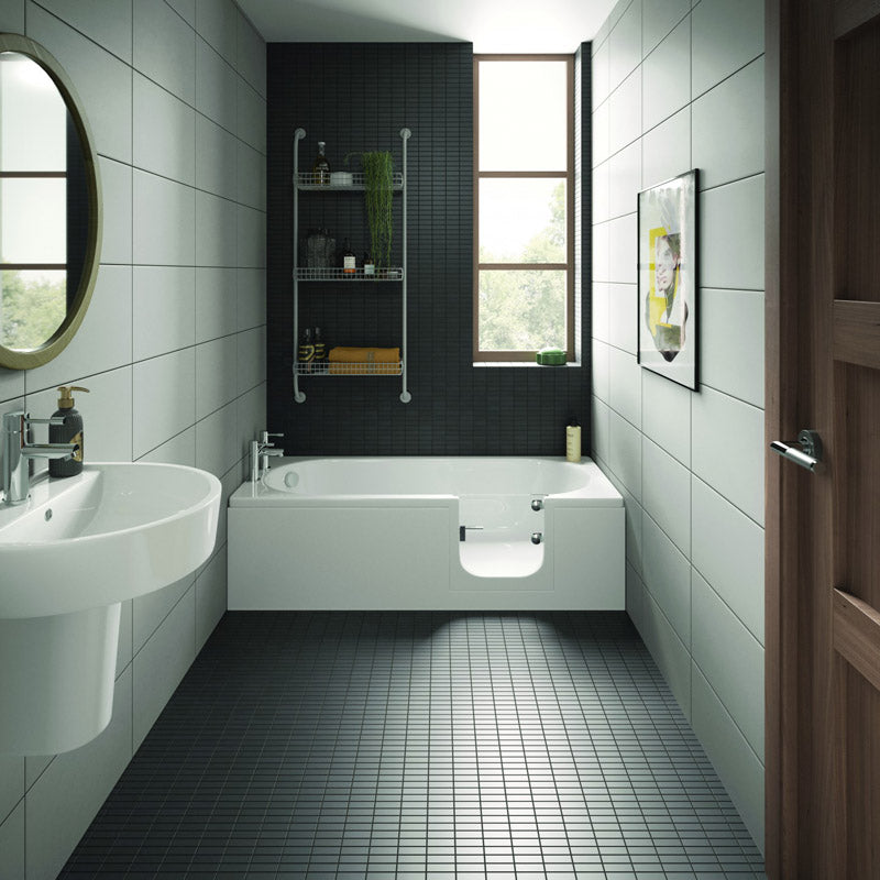 A modern bathroom interior with a white bathtub set into a dark tiled wall, white wall-mounted sink, black floor tiles, a round mirror, shelving, and a window.