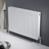A white panel radiator mounted on a dark gray wall, supported by two white wall anchors, with a white tiled floor and a decorative metallic bowl in the foreground.