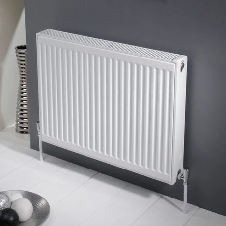 A white panel radiator mounted on a dark gray wall, supported by two white wall anchors, with a white tiled floor and a decorative metallic bowl in the foreground.