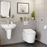 A modern bathroom scene showing a white wall-mounted toilet and a white pedestal sink with a chrome faucet, set against gray tiled walls. A black-framed wall mirror, framed artwork, small potted plants, and a toilet brush are visible.