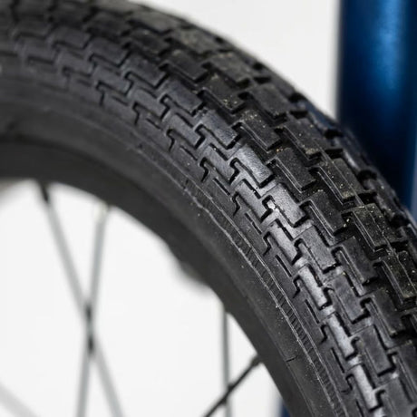 Close-up of a black wheelchair tyre with a zigzag tread pattern, mounted on a wheel with thin silver spokes, against a white background with a blue frame element visible on the right.
