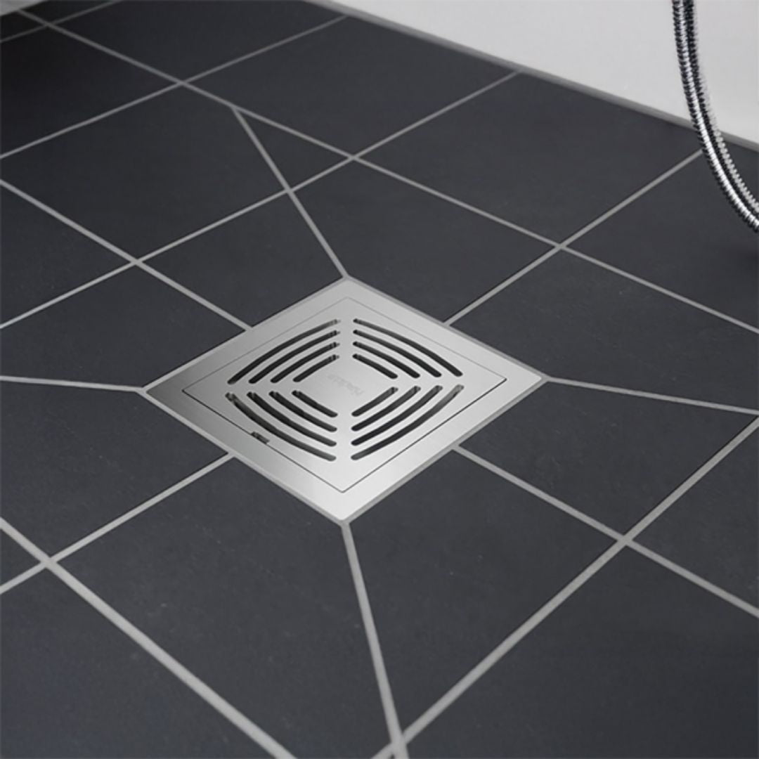 A square stainless steel floor drain with a slotted center, installed on a dark gray tiled floor with white grout lines.