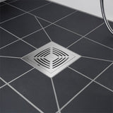 A square stainless steel floor drain with a slotted center, installed on a dark gray tiled floor with white grout lines.