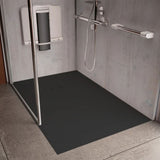 A black rectangular shower floor mat installed in a modern gray tiled shower stall with a glass door, chrome grab handles, and a wall-mounted shower seat.